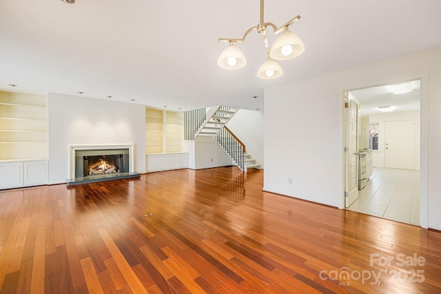 unfurnished living room featuring built in shelves, a notable chandelier, a glass covered fireplace, hardwood / wood-style floors, and stairs