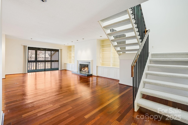 unfurnished living room with baseboards, stairway, built in features, a lit fireplace, and hardwood / wood-style floors