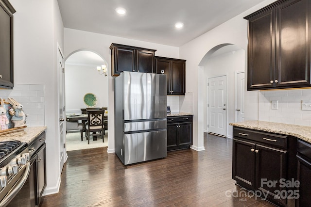 kitchen featuring arched walkways, stainless steel appliances, dark wood-type flooring, dark brown cabinetry, and light stone countertops