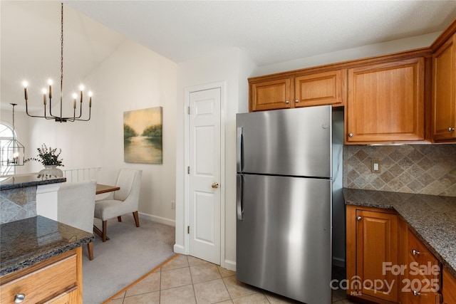 kitchen with light tile patterned floors, freestanding refrigerator, brown cabinets, dark stone counters, and tasteful backsplash