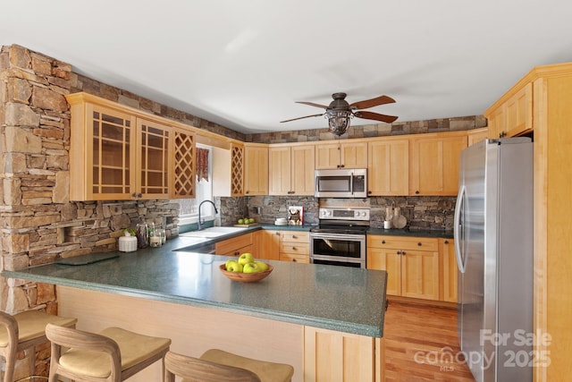 kitchen with dark countertops, appliances with stainless steel finishes, a peninsula, light brown cabinetry, and a sink