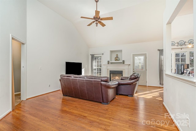 living room with a ceiling fan, baseboards, high vaulted ceiling, light wood-style flooring, and a lit fireplace