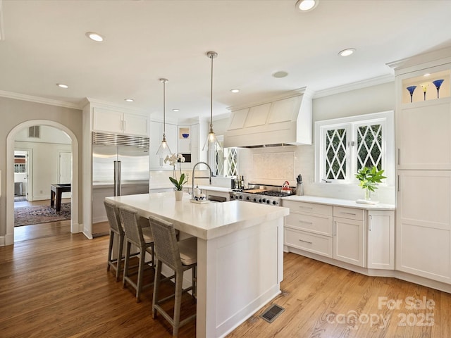 kitchen with arched walkways, built in refrigerator, a kitchen island with sink, custom exhaust hood, and light countertops