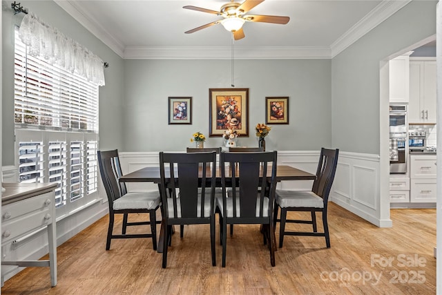 dining space featuring a wainscoted wall, light wood finished floors, a ceiling fan, and crown molding