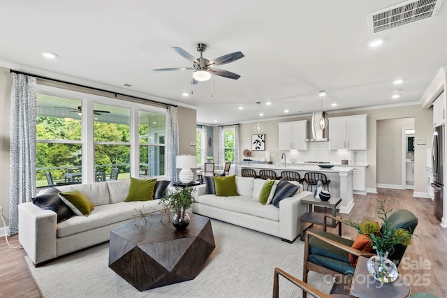 living area with light wood finished floors, visible vents, a ceiling fan, crown molding, and recessed lighting