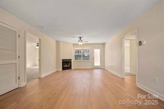 unfurnished living room with a ceiling fan, baseboards, light wood finished floors, and a lit fireplace