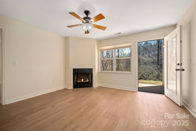 unfurnished living room featuring visible vents, baseboards, a warm lit fireplace, ceiling fan, and light wood-style floors