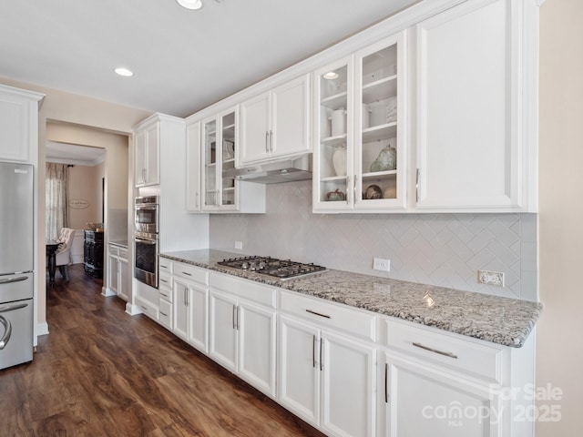 kitchen with glass insert cabinets, light stone countertops, under cabinet range hood, white cabinets, and stainless steel appliances