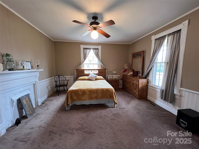 carpeted bedroom featuring ceiling fan, a fireplace, visible vents, ornamental molding, and wainscoting