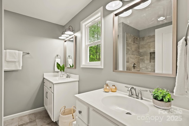 bathroom featuring a tile shower, baseboards, two vanities, and a sink