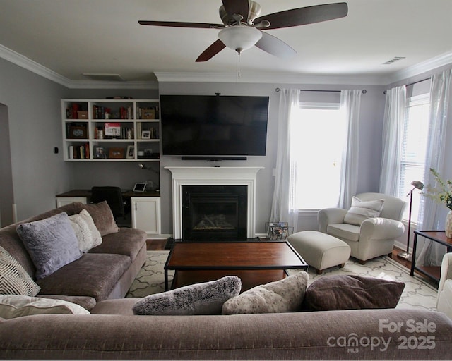 living room featuring ceiling fan, wood finished floors, visible vents, ornamental molding, and a glass covered fireplace