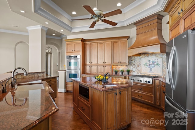 kitchen with a raised ceiling, an island with sink, dark stone countertops, stainless steel appliances, and a sink