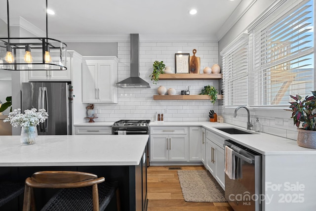 kitchen with wall chimney range hood, light countertops, ornamental molding, appliances with stainless steel finishes, and a sink