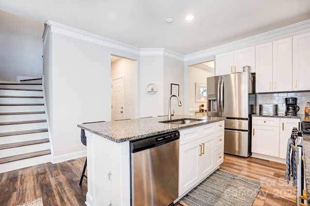 kitchen with a sink, ornamental molding, appliances with stainless steel finishes, light stone countertops, and a center island with sink