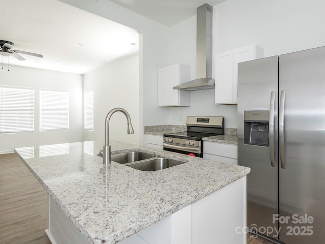 kitchen featuring a center island with sink, appliances with stainless steel finishes, white cabinets, a sink, and wall chimney range hood