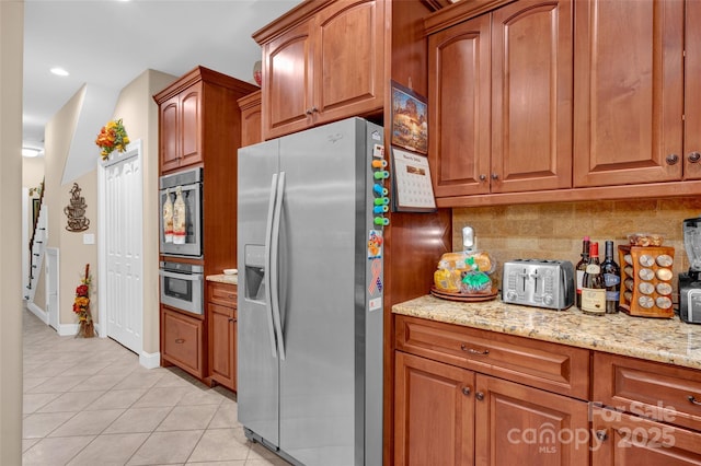 kitchen with light tile patterned floors, light stone countertops, stainless steel appliances, tasteful backsplash, and brown cabinets