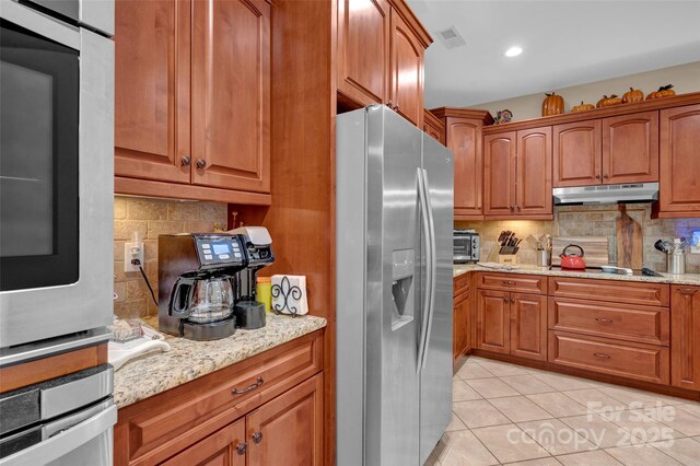 kitchen featuring under cabinet range hood, light stone counters, backsplash, appliances with stainless steel finishes, and light tile patterned floors