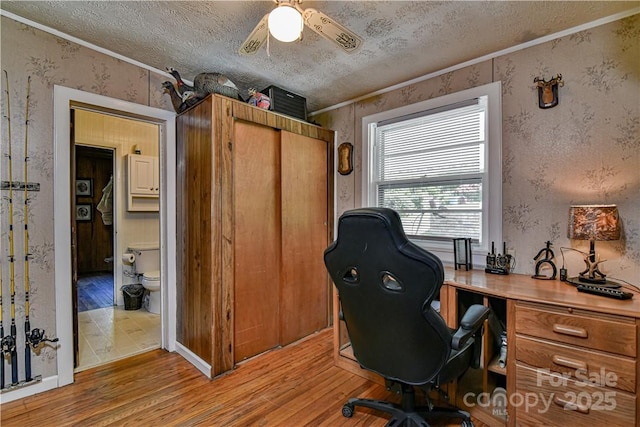 home office featuring light wood-style flooring, ornamental molding, a textured ceiling, wallpapered walls, and ceiling fan