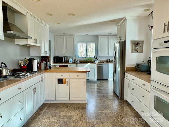 kitchen with stainless steel appliances, white cabinetry, a sink, wall chimney range hood, and a peninsula