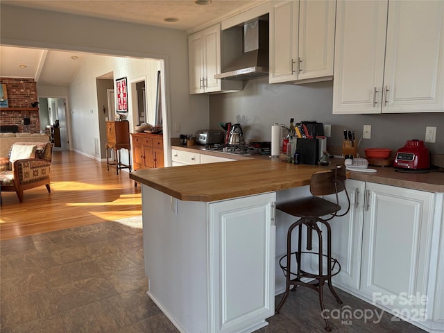 kitchen featuring wood counters, white cabinetry, wall chimney exhaust hood, a kitchen bar, and stainless steel gas stovetop