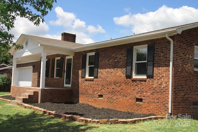 view of side of home with covered porch, an attached garage, crawl space, brick siding, and a chimney