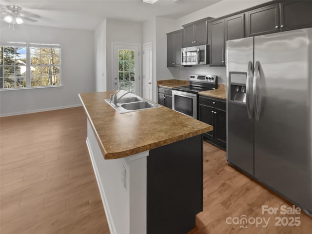 kitchen with light wood-style flooring, a sink, baseboards, appliances with stainless steel finishes, and an island with sink