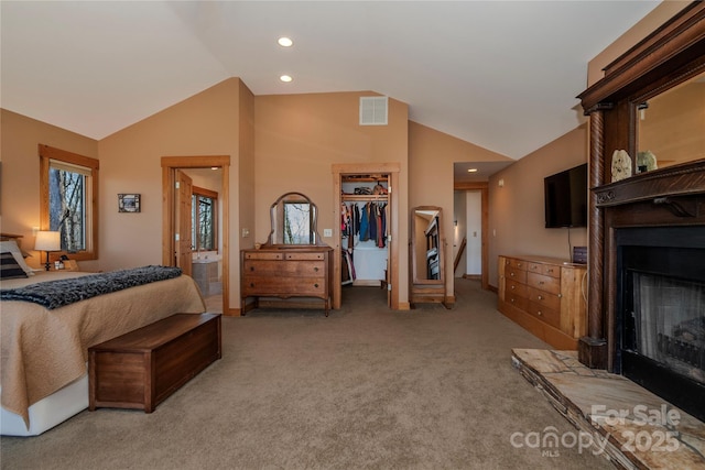 bedroom with visible vents, a fireplace with raised hearth, a walk in closet, light colored carpet, and vaulted ceiling