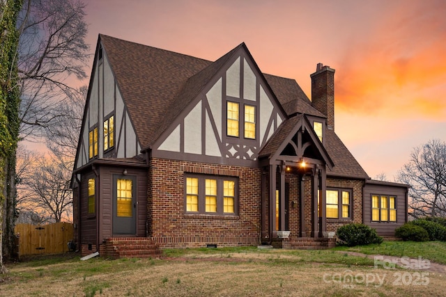 tudor-style house featuring brick siding, fence, roof with shingles, stucco siding, and a chimney