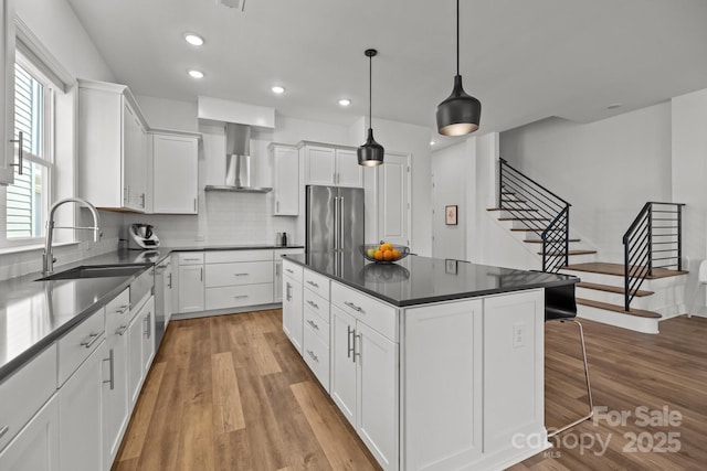 kitchen with stainless steel appliances, dark countertops, a sink, wood finished floors, and wall chimney exhaust hood