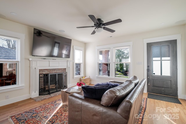 living area featuring baseboards, light wood-type flooring, ceiling fan, and a brick fireplace
