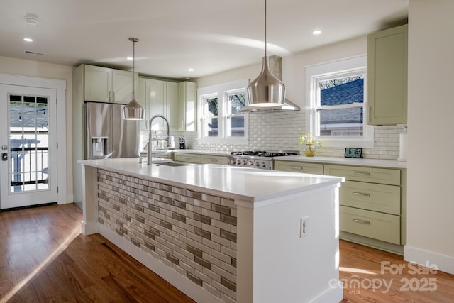 kitchen with light countertops, island range hood, stainless steel appliances, a kitchen island with sink, and wood finished floors