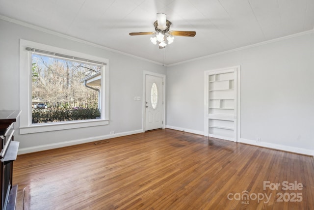 unfurnished living room featuring built in shelves, wood finished floors, crown molding, baseboards, and ceiling fan