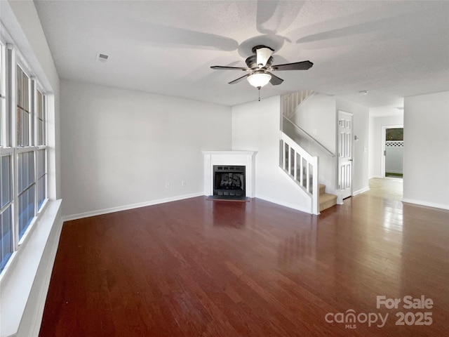 unfurnished living room featuring baseboards, a fireplace with flush hearth, ceiling fan, wood finished floors, and stairs