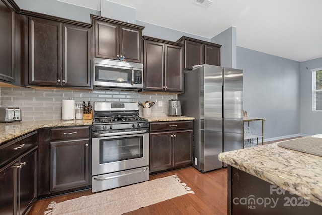 kitchen featuring dark brown cabinetry, decorative backsplash, light stone counters, wood finished floors, and stainless steel appliances