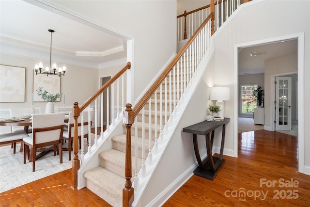 stairway with wood finished floors, baseboards, ornamental molding, a raised ceiling, and a chandelier