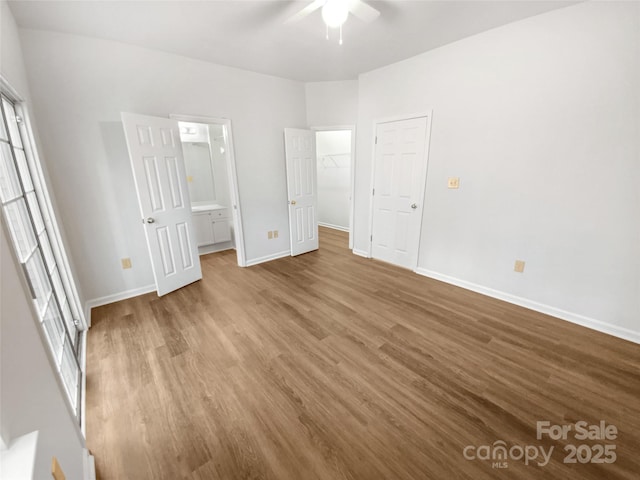 unfurnished bedroom with a walk in closet, ensuite bathroom, a ceiling fan, wood finished floors, and baseboards