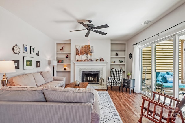 living room with built in shelves, a brick fireplace, wood finished floors, and a ceiling fan