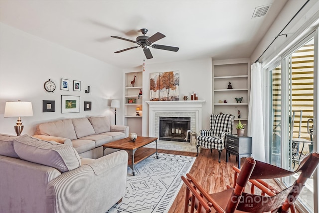 living area with visible vents, ceiling fan, wood finished floors, a brick fireplace, and built in shelves