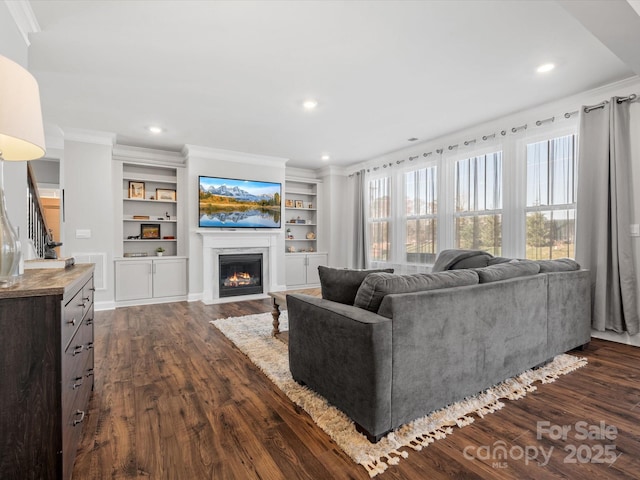 living area with built in shelves, crown molding, dark wood finished floors, a fireplace with flush hearth, and stairs