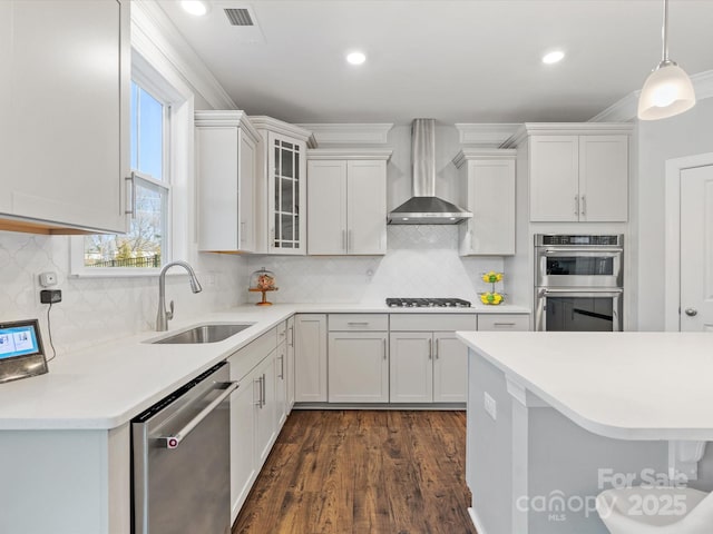 kitchen with a sink, hanging light fixtures, light countertops, stainless steel appliances, and wall chimney exhaust hood