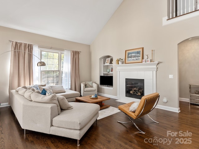 living area featuring baseboards, a tiled fireplace, built in features, arched walkways, and dark wood-style flooring