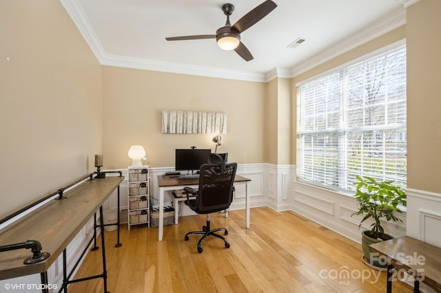 home office featuring a wainscoted wall, wood finished floors, a ceiling fan, visible vents, and crown molding