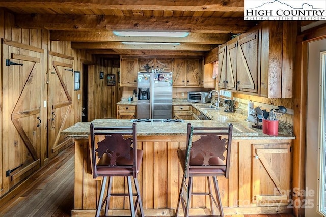 kitchen featuring beamed ceiling, a peninsula, wood walls, stainless steel refrigerator with ice dispenser, and a sink