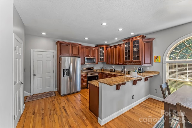 kitchen with stone counters, a peninsula, light wood-style flooring, appliances with stainless steel finishes, and a kitchen bar