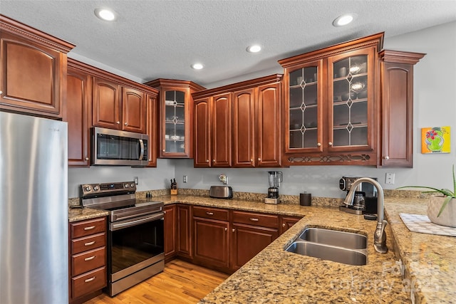 kitchen featuring a sink, stainless steel appliances, light wood finished floors, glass insert cabinets, and light stone countertops