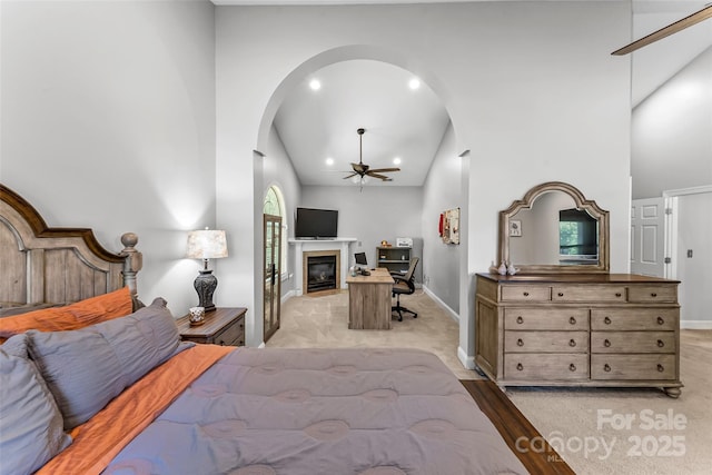 bedroom with baseboards, light colored carpet, arched walkways, a glass covered fireplace, and high vaulted ceiling