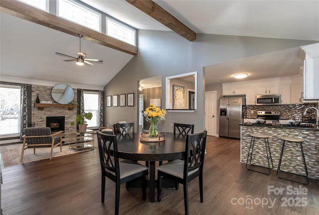 dining room featuring a ceiling fan, high vaulted ceiling, a fireplace, dark wood-type flooring, and beamed ceiling