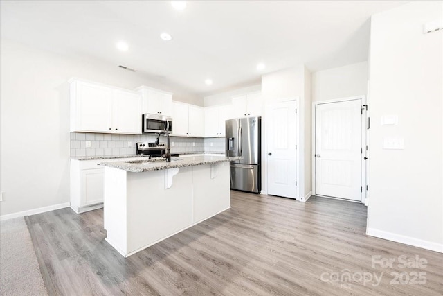 kitchen featuring a kitchen island with sink, stainless steel appliances, white cabinetry, decorative backsplash, and light stone countertops