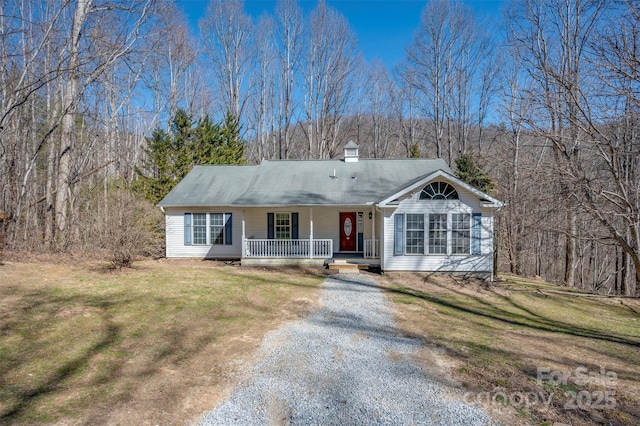 view of front of house featuring a chimney, a porch, gravel driveway, a front lawn, and a wooded view