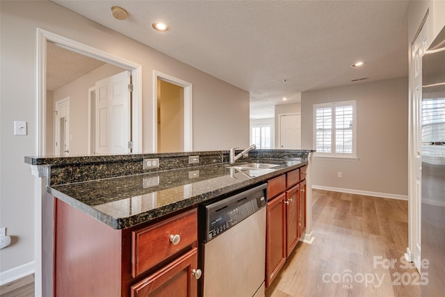 kitchen featuring light wood finished floors, an island with sink, dark stone countertops, stainless steel dishwasher, and a sink